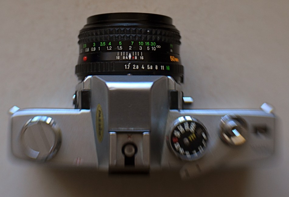 My antedeluvian Minolta SRT-500 camera body, crowned by its original f/1.7 50mm prime kit lens. Guess which one is worth its weight in gold? 